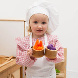 Child in a chef's outfit holding wooden bowls with colorful play food in a kitchen setting